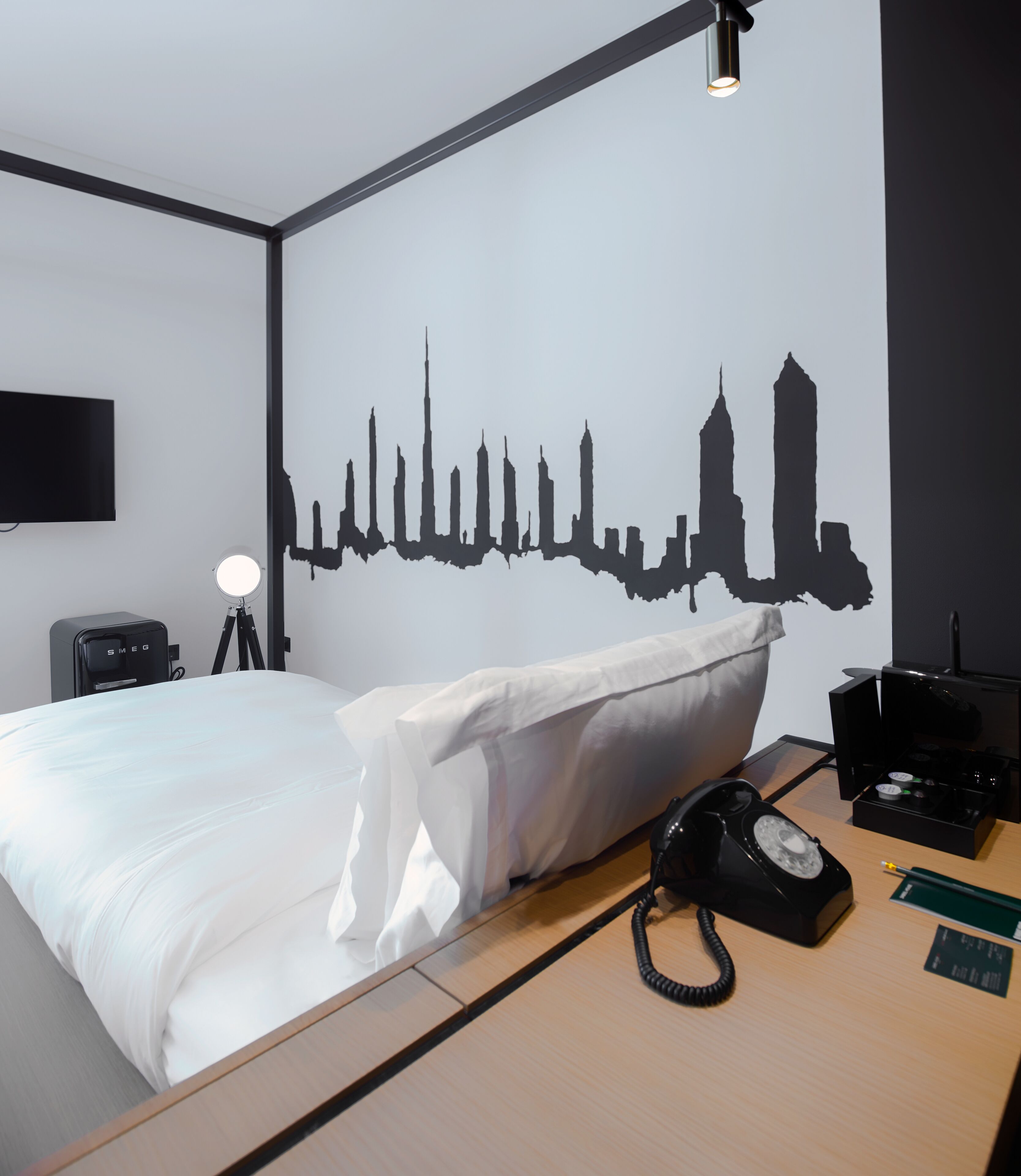 A hotel room with a bed, a desk with a telephone, a lamp, and a wall with a city skyline silhouette.