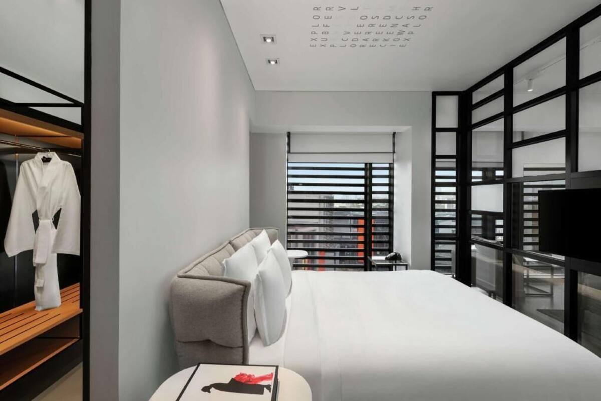 A modern hotel room with a bed, a bedside table, a robe hanging, and a large window with blinds.