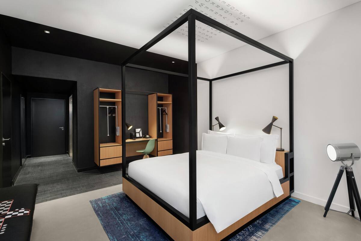 A modern bedroom with a four-poster bed, a desk with a chair, a wardrobe, and a telescope.
