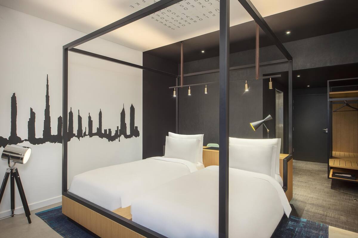 A modern hotel room with a large bed, a city skyline mural, a desk with a lamp, and a wardrobe.