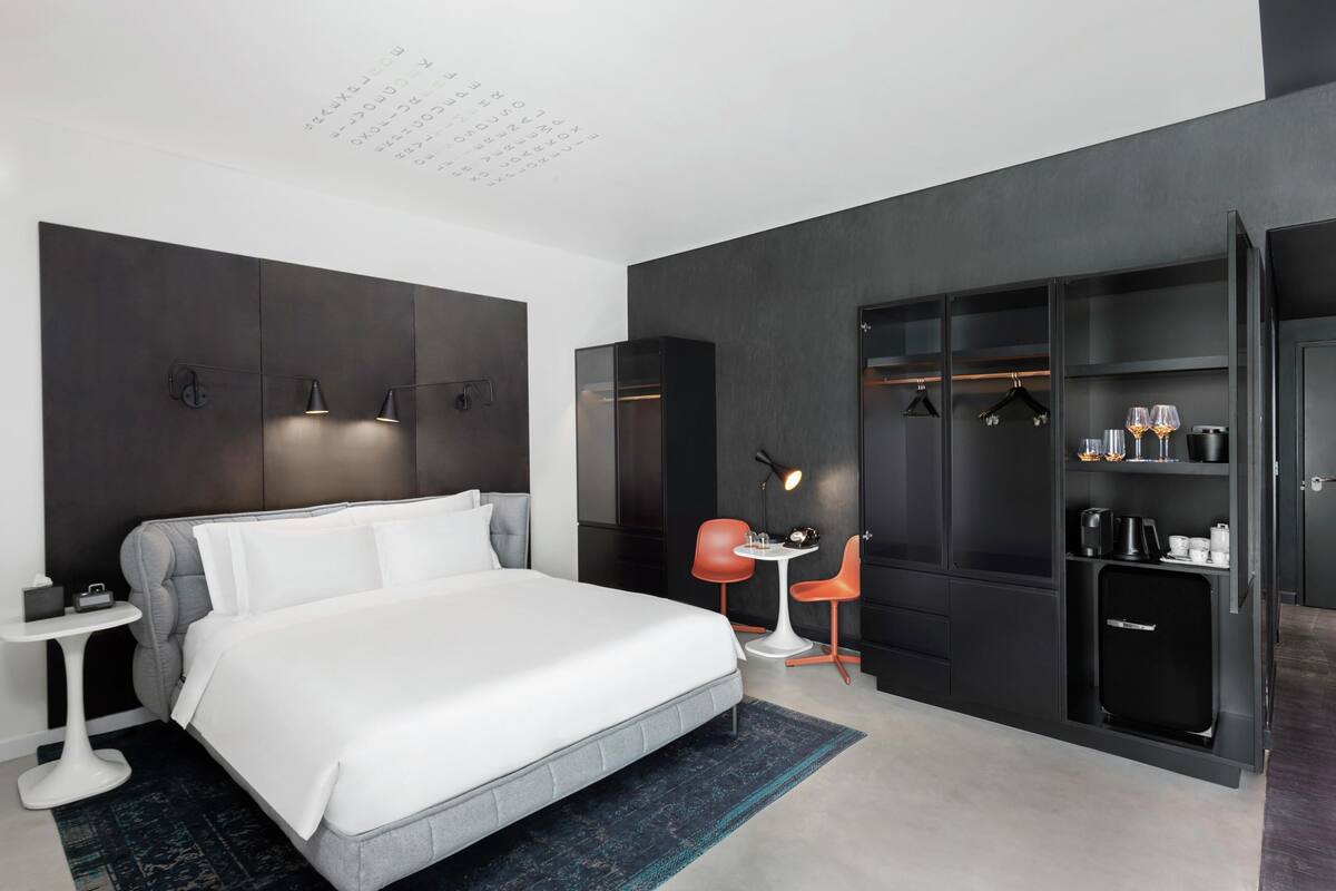 A modern hotel room with a large bed, a small table, two chairs, a wardrobe, and a desk with a lamp.
