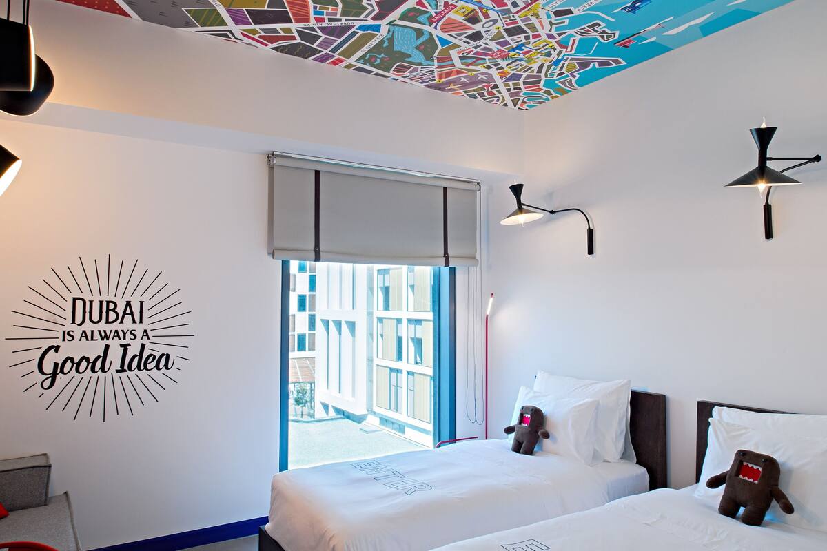 Hotellihuone, jossa on kaksi sänkyä, katossa kaupunkimaisemaa esittävä seinämaalaus, ikkuna, josta avautuu näkymä rakennuksiin, ja seinässä oleva kyltti, jossa lukee: ”DUBAI ON AINA HYVÄ VALINTA”.