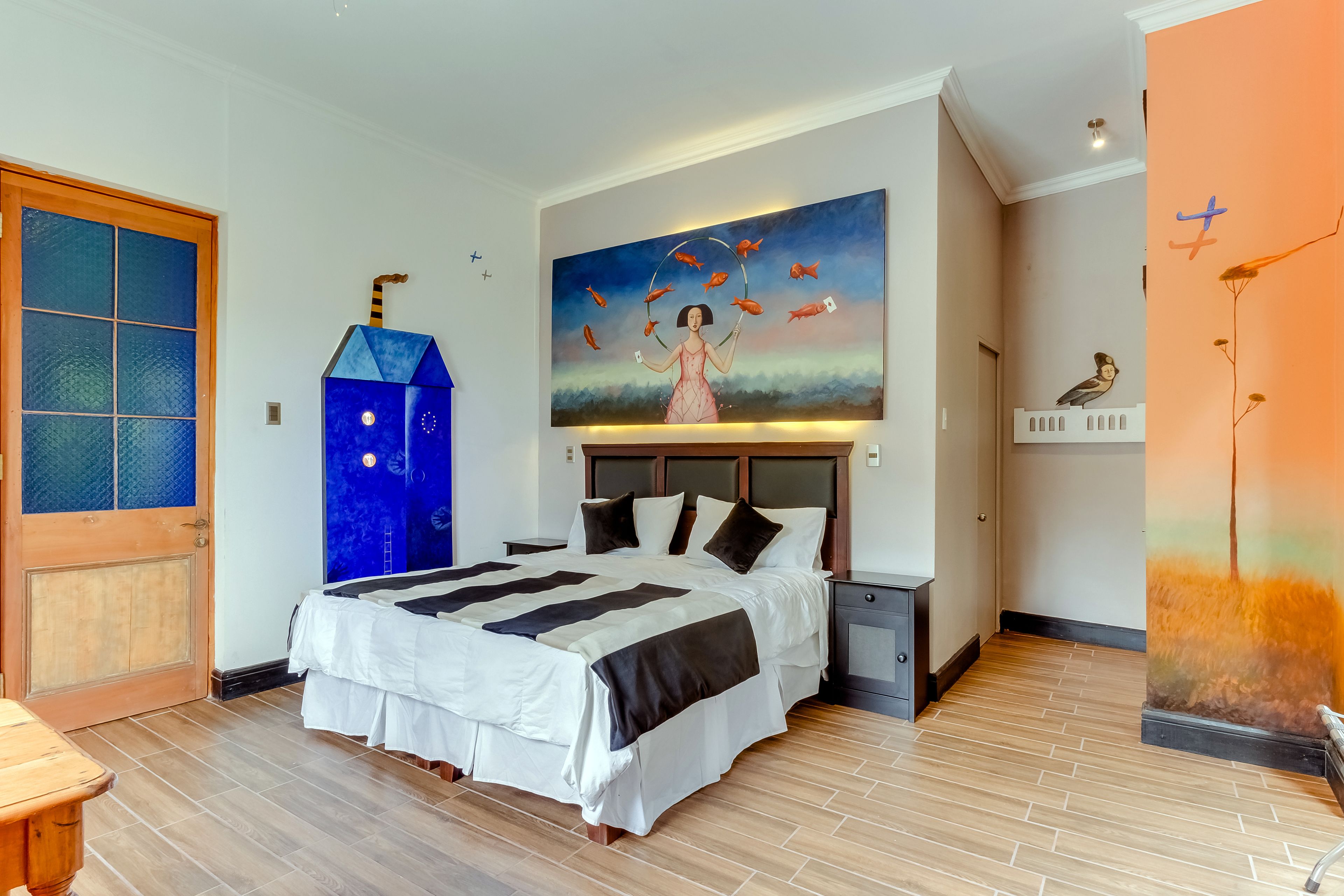 Une chambre à coucher avec un grand lit, un tableau représentant une femme et des poissons, une porte en bois et une table de chevet.