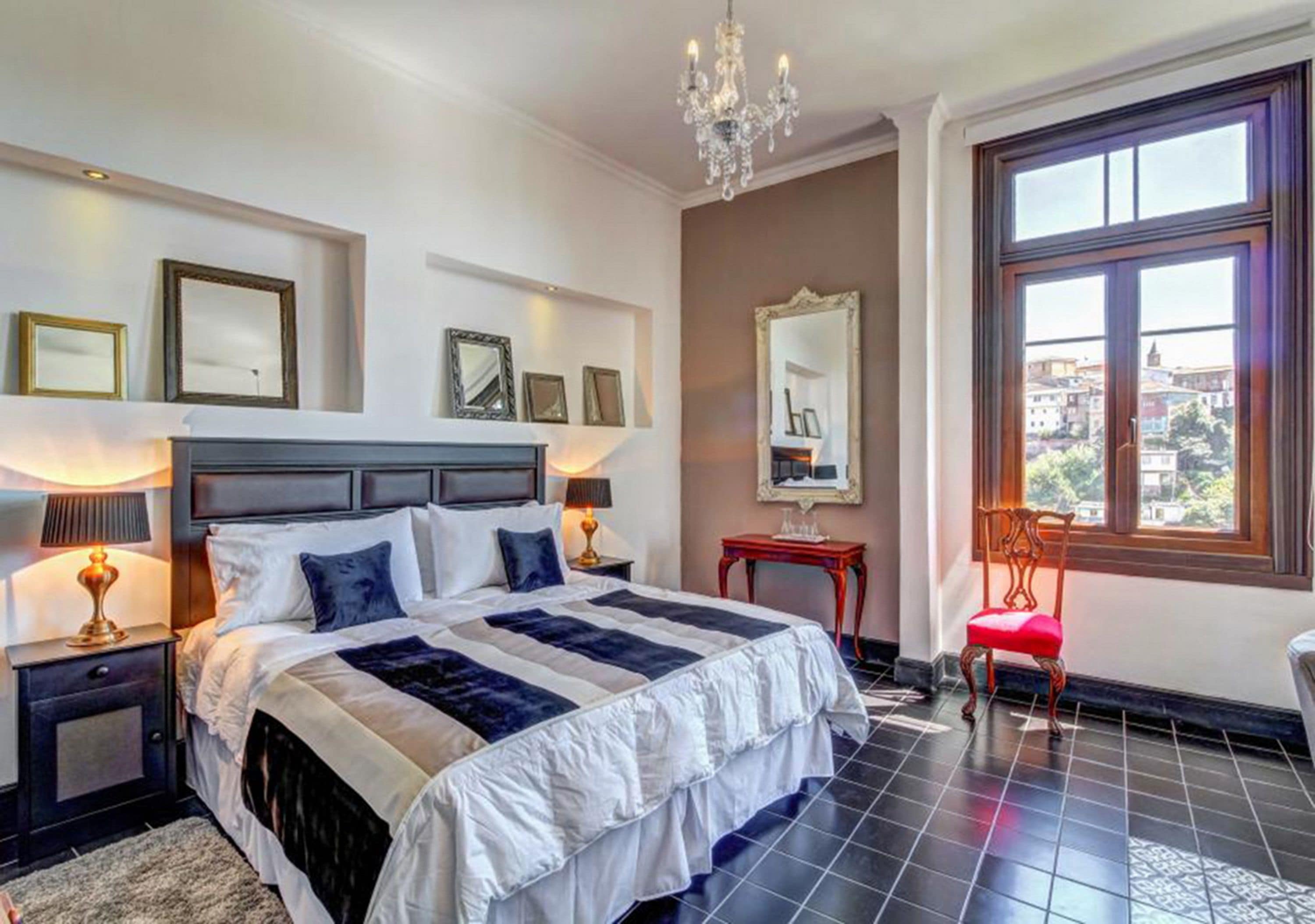 A neatly arranged bedroom with a large bed, bedside lamps, a mirror, and a view through a window.