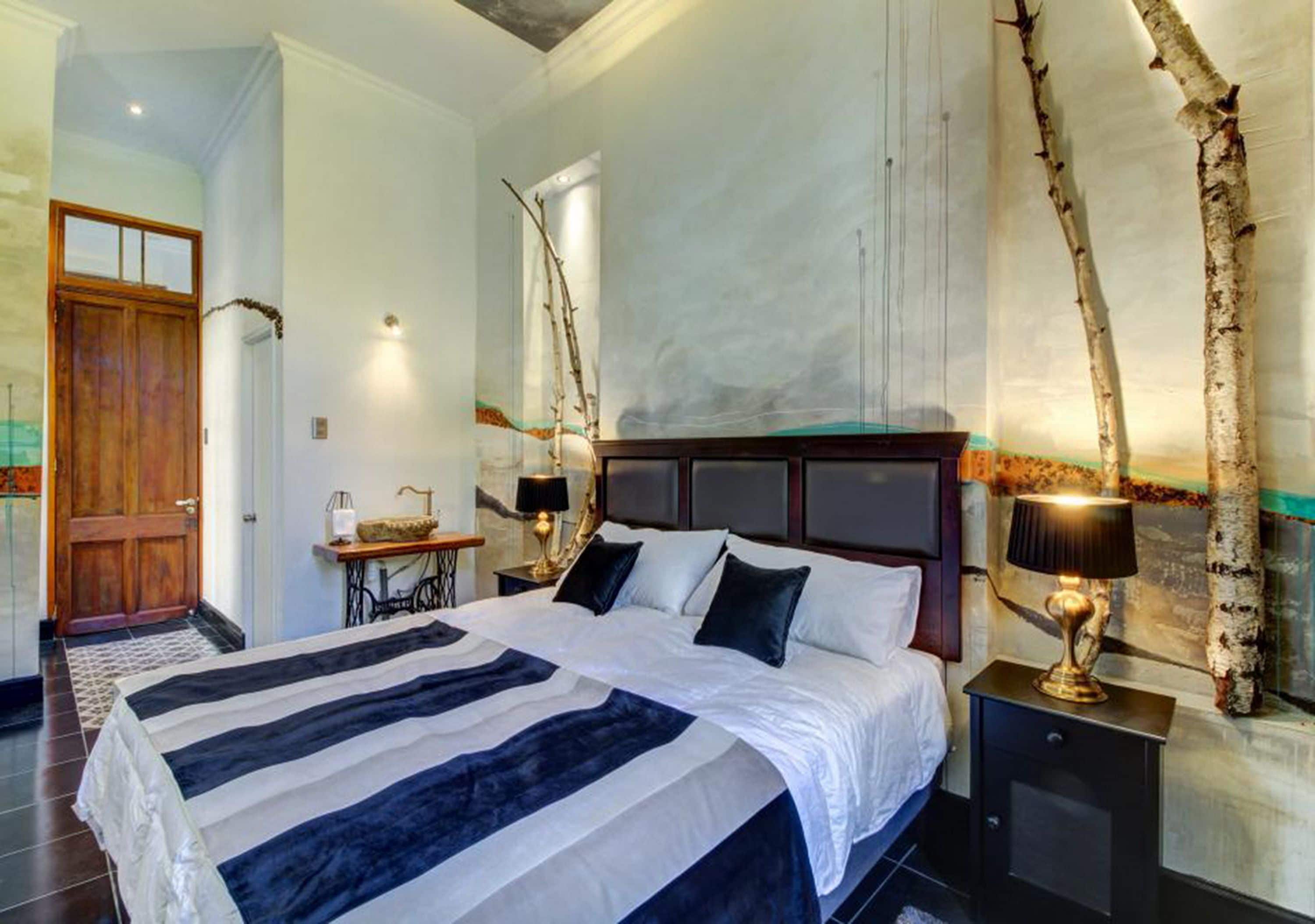A hotel room with a large bed, a wooden door, a sewing machine, and a wall-mounted lamp.