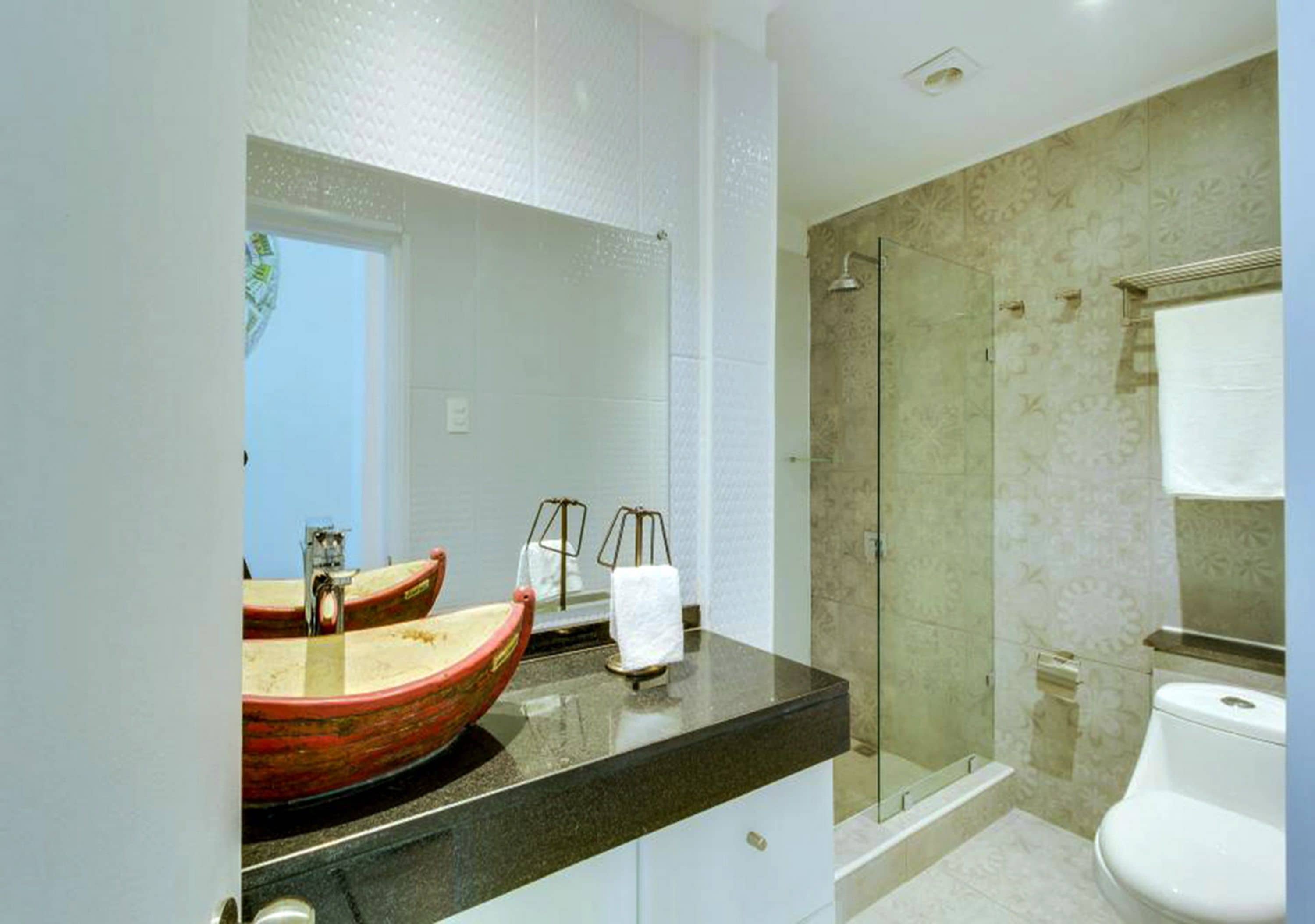 A bathroom with a large mirror, a red bowl sink, a glass shower enclosure, and a white toilet.