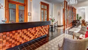 Vestíbulo de hotel con mostrador de recepción, baldosas de suelo estampadas y un área de descanso con sillas y una mesa.
