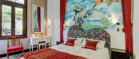Un dormitorio con cabecera de estampado de cebra, una cama grande con ropa de cama roja y blanca, una cortina roja, un tocador blanco, un espejo y una ventana con vistas a árboles.