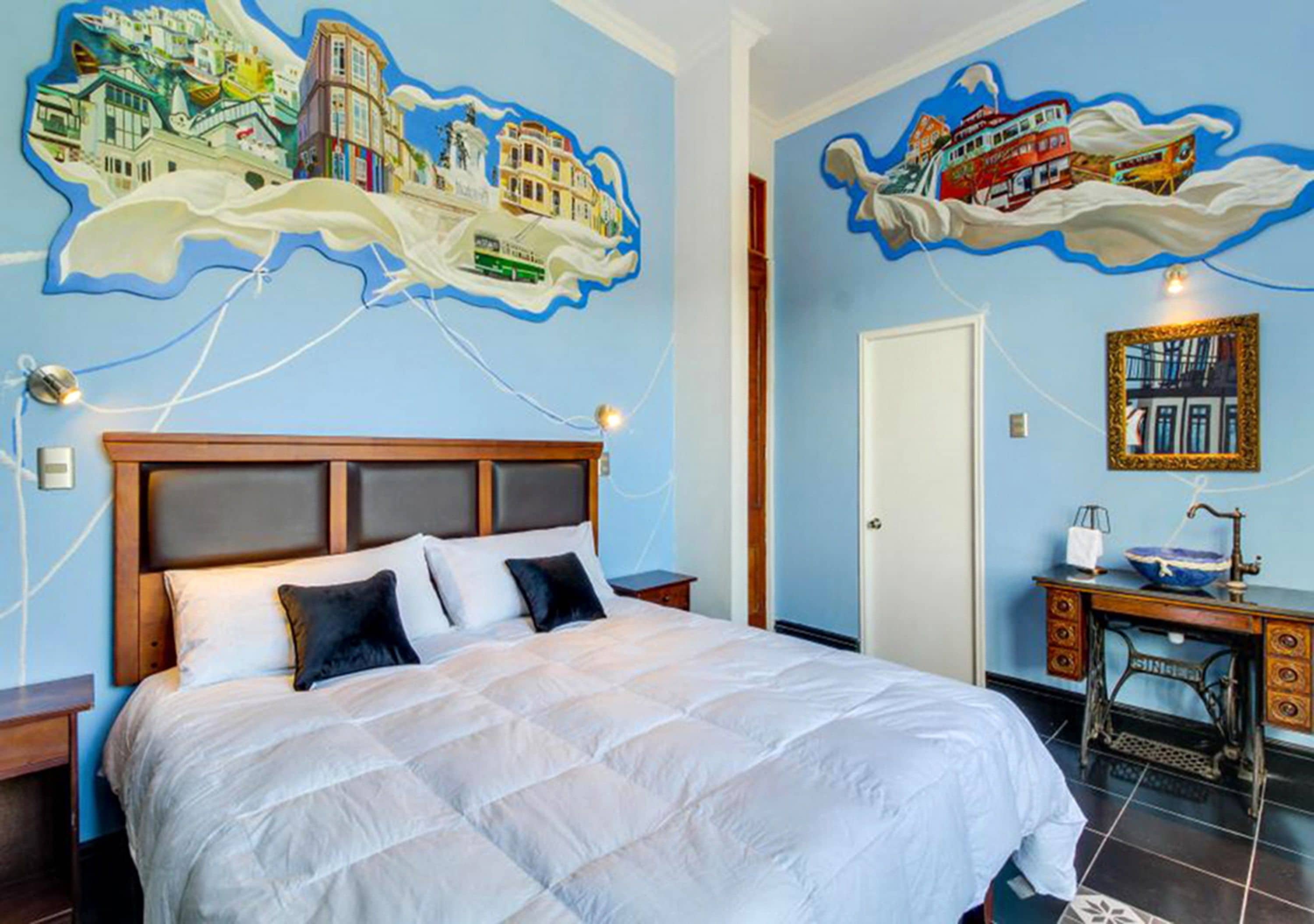 Une chambre d’hôtel avec un grand lit, un bureau sur lequel se trouve une machine à coudre ancienne, et un mur orné d’une fresque représentant un paysage urbain.