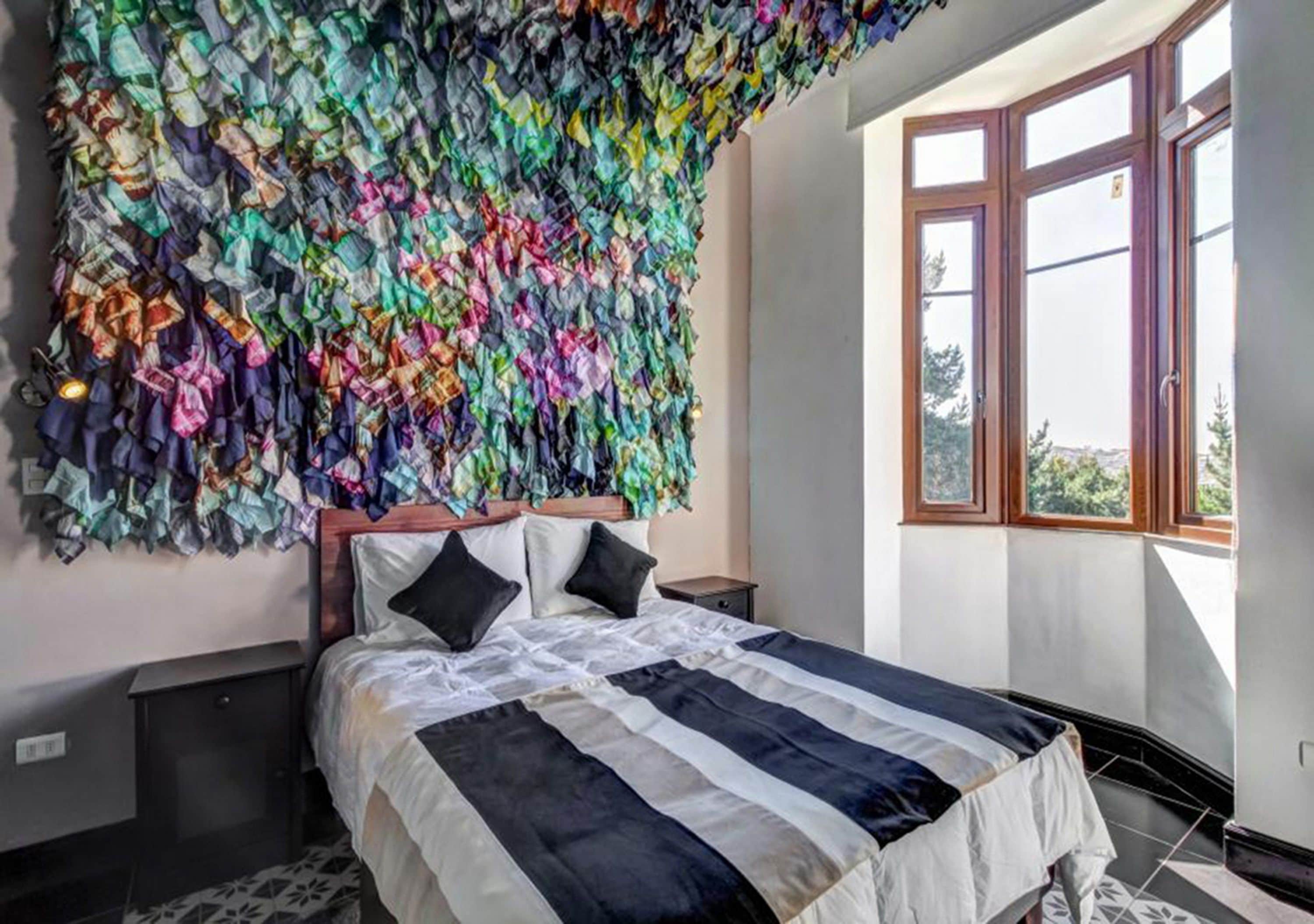 A bedroom with a bed, pillows, a nightstand, a window, and a wall covered with colorful fabric.
