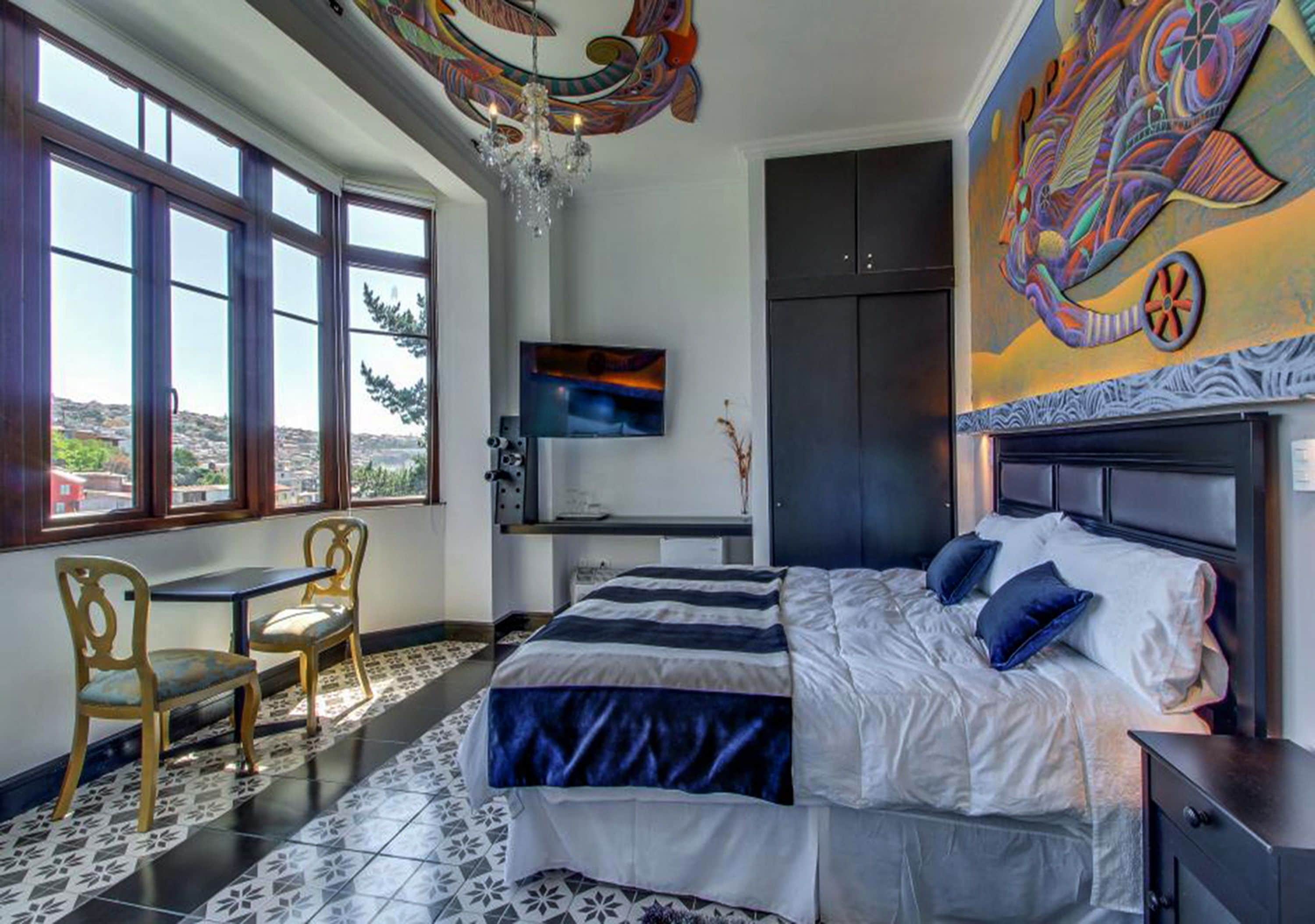 A bedroom with a bed, a desk, and a TV. There are windows and a view of the outside.