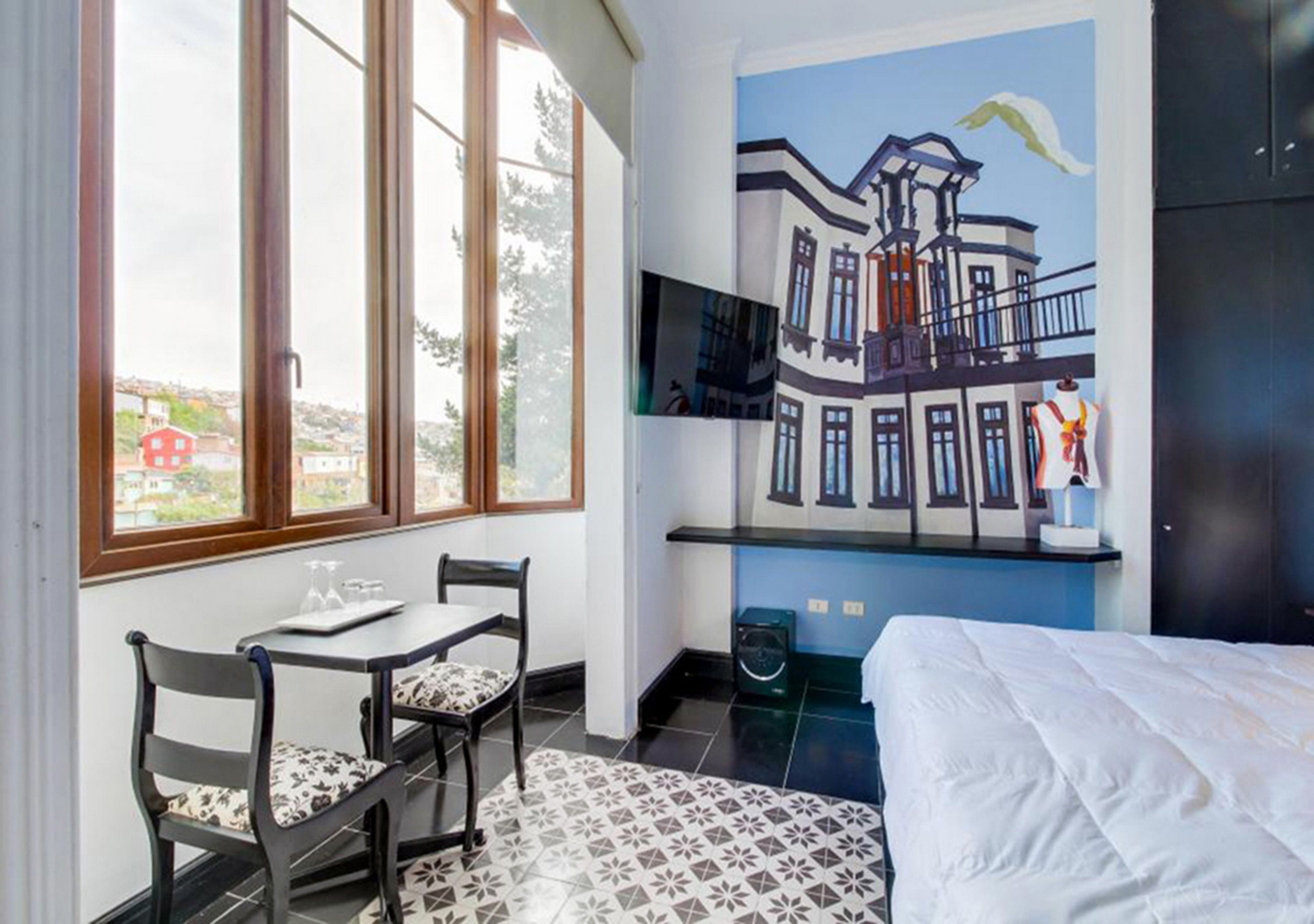 A hotel room with a bed, a dining table with chairs, a mural on the wall, and a view of the outside.
