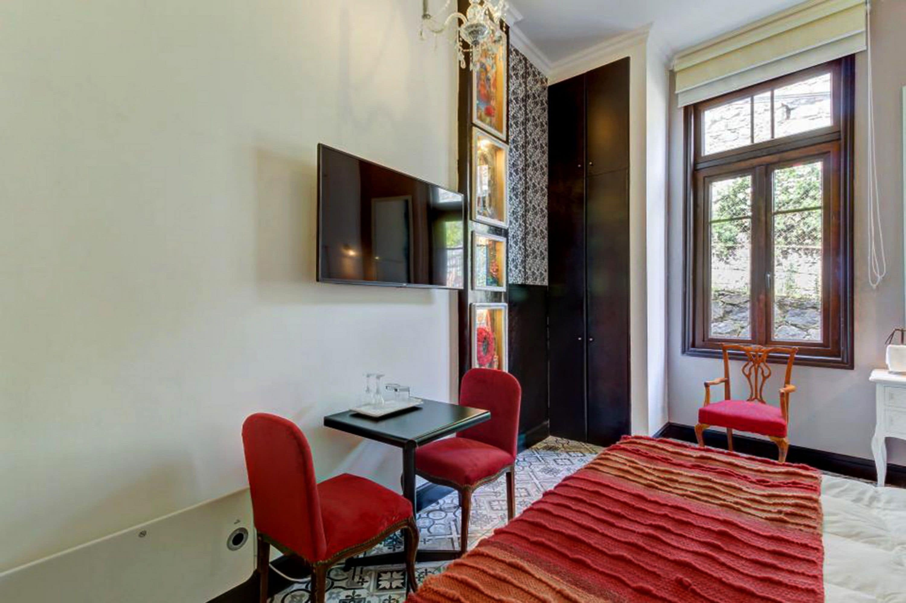 A hotel room with a bed, a desk with two red chairs, a TV mounted on the wall, and a window with a view of greenery.