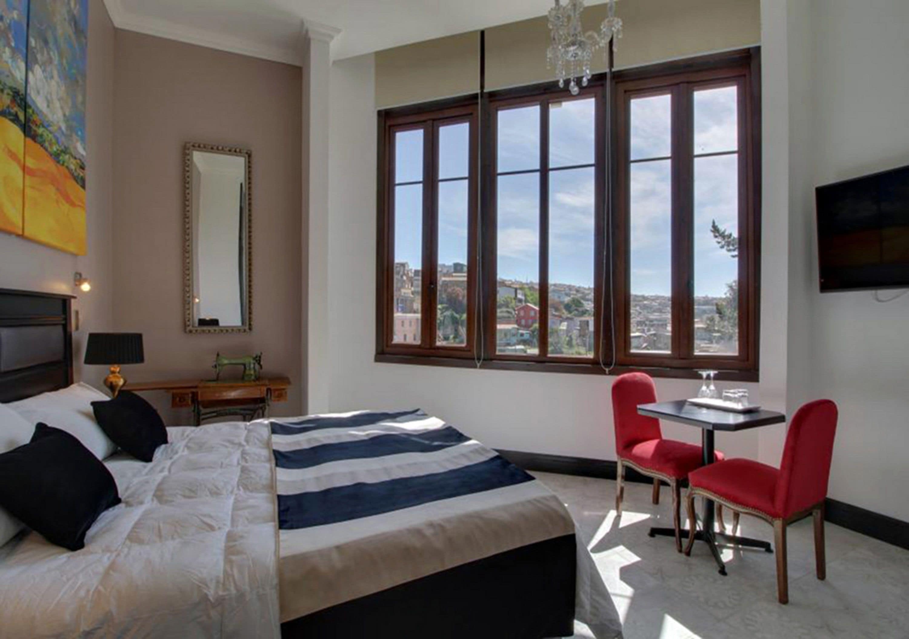 A bedroom with a large bed, a desk, two red chairs, a TV, and a city view through large windows.