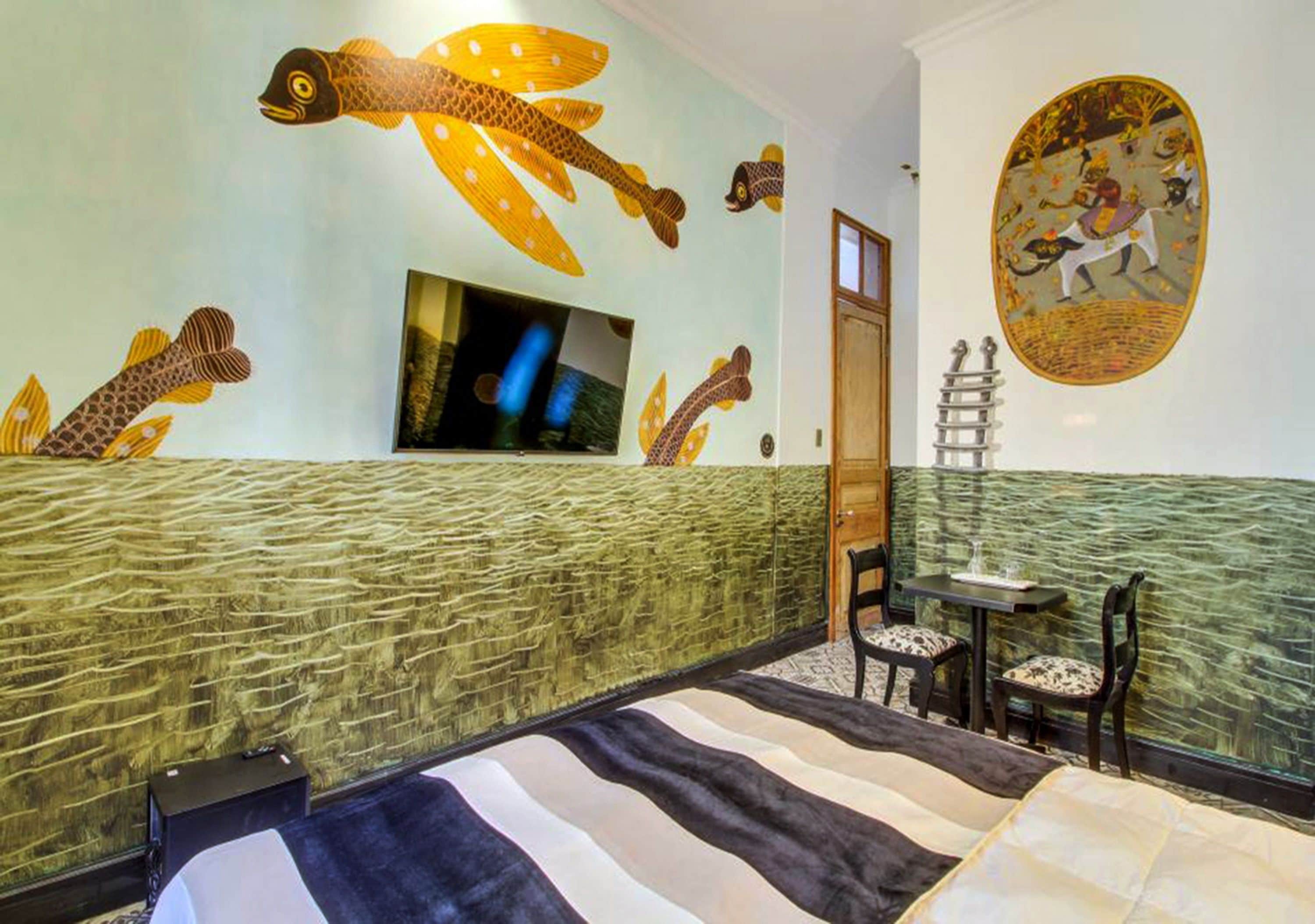 A hotel room with a bed, a TV, a desk with chairs, and walls decorated with fish and a mural.