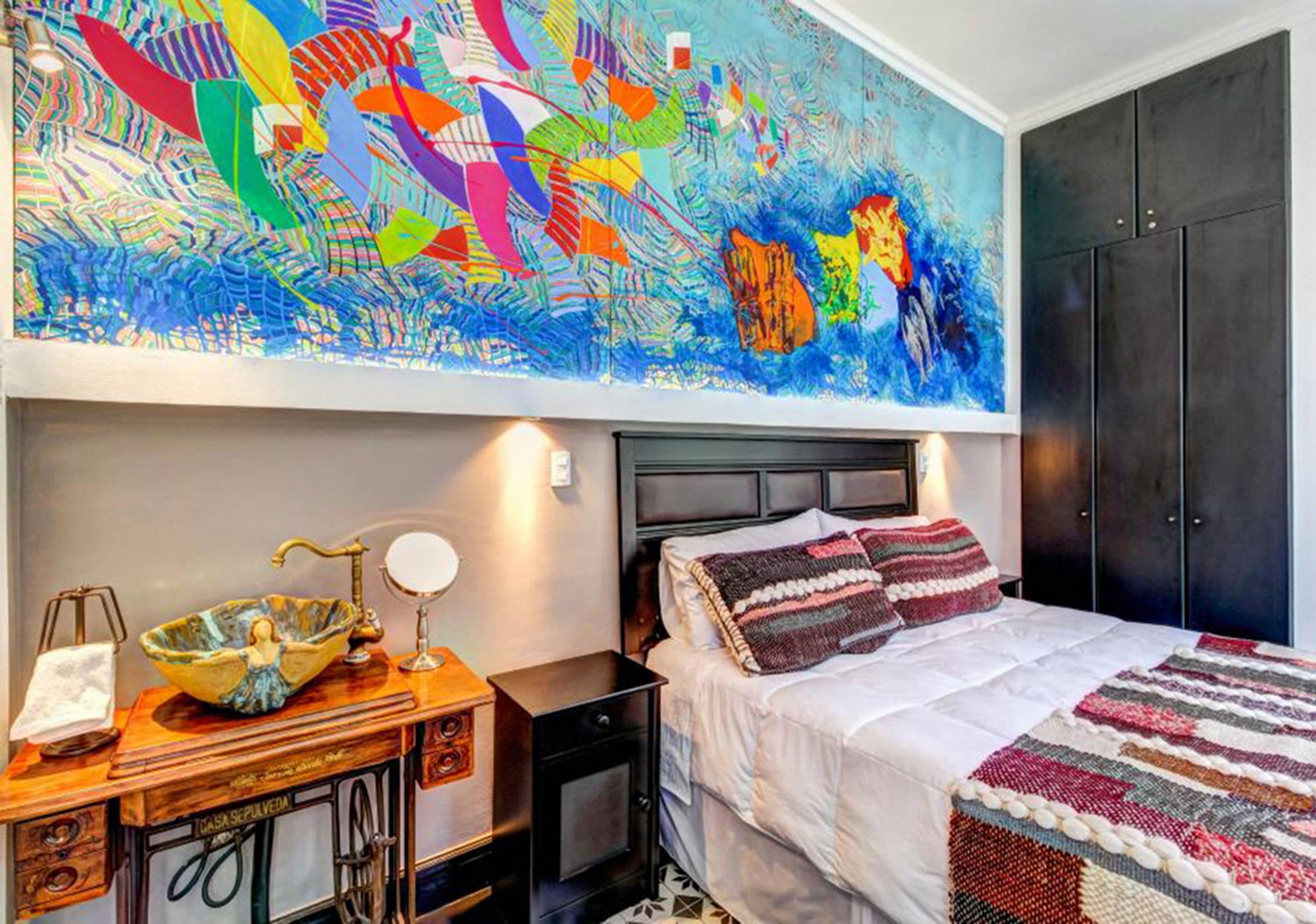 Un dormitorio con un cuadro abstracto grande, una cama con almohadillas a rayas, una mesita de noche de madera con una lámpara y un cuenco decorativo.