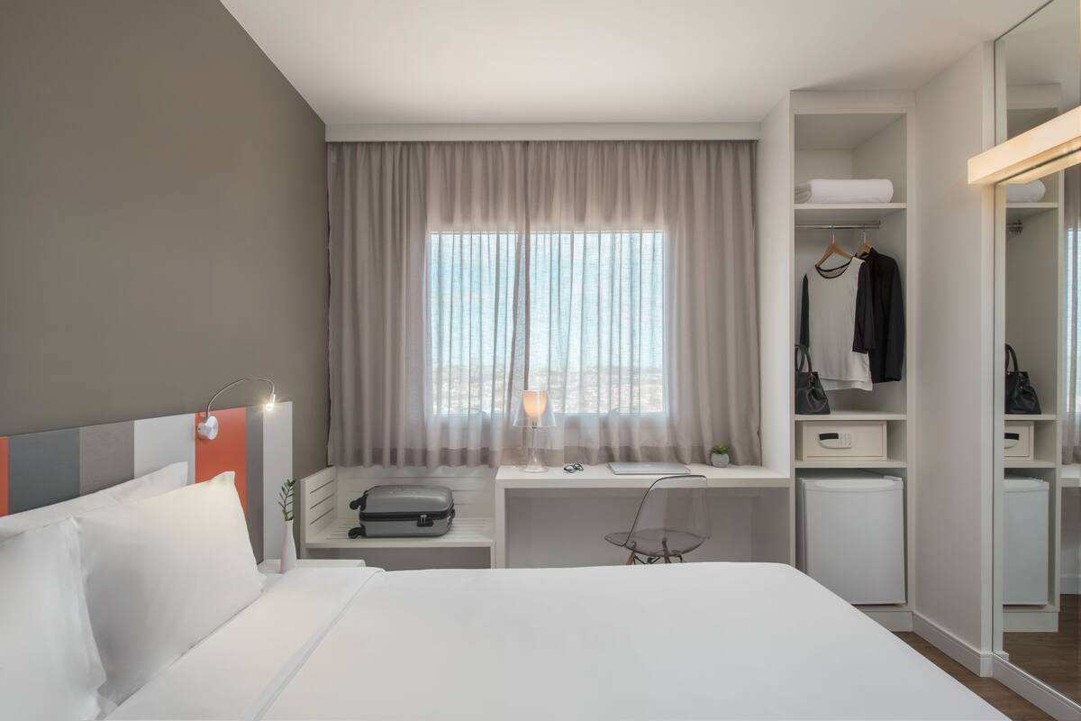 A modern hotel room with a large bed, a desk with a lamp, a wardrobe, and a view of the outdoors through sheer curtains.
