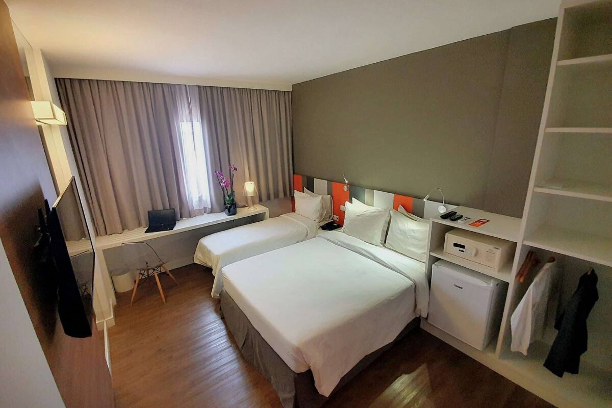 A hotel room with two beds, a desk, a chair, a small table with a lamp, a wardrobe, and a window with curtains.