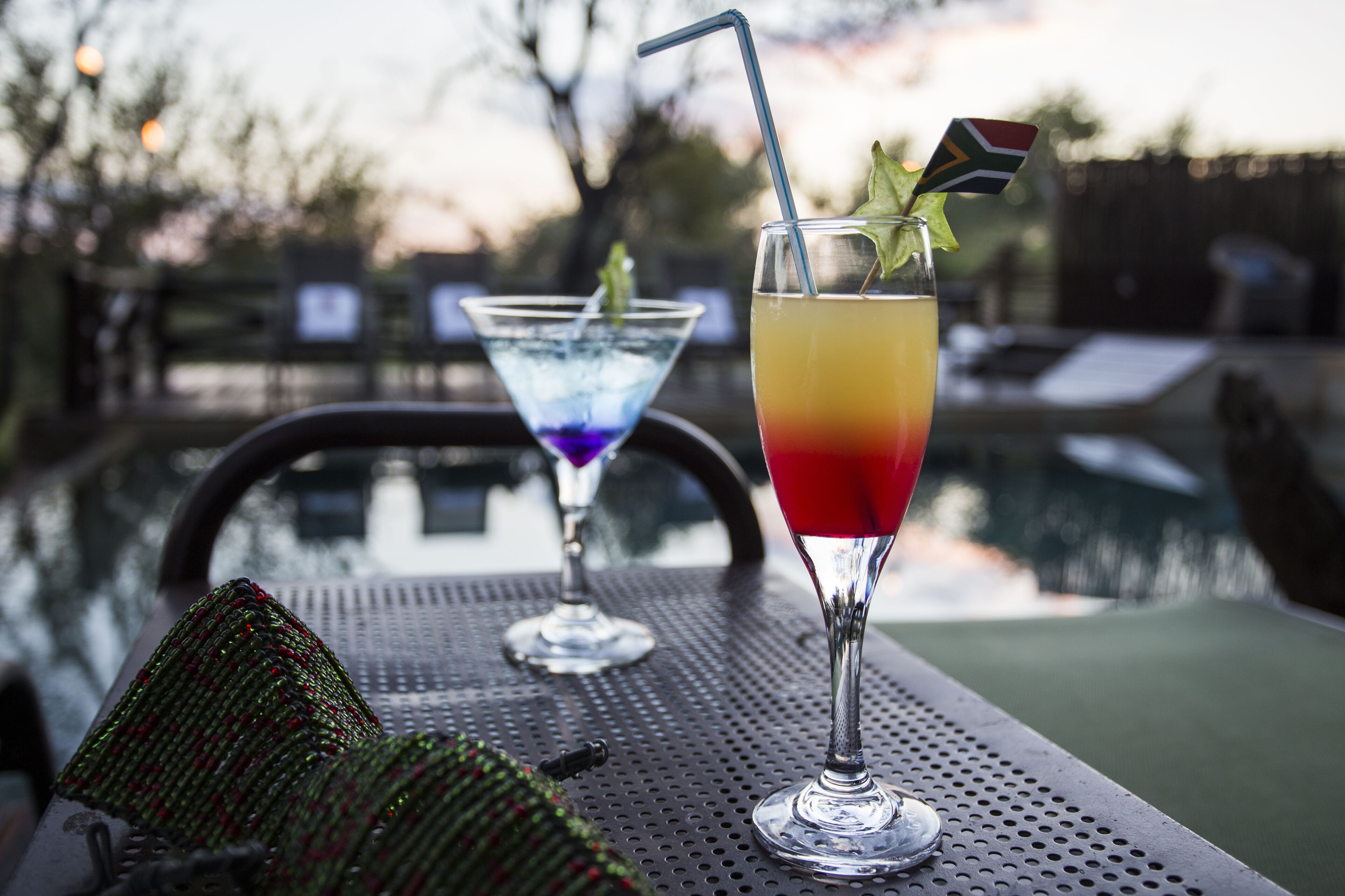 Two cocktails on a metal table near a pool, with a chair and a decorative item in the foreground.
