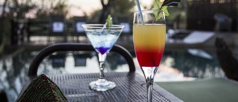 Two cocktails on a metal table near a pool, with a chair and a decorative item in the foreground.
