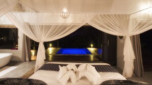 A bedroom with a large bed, a bathtub, and a view of a pool at night.
