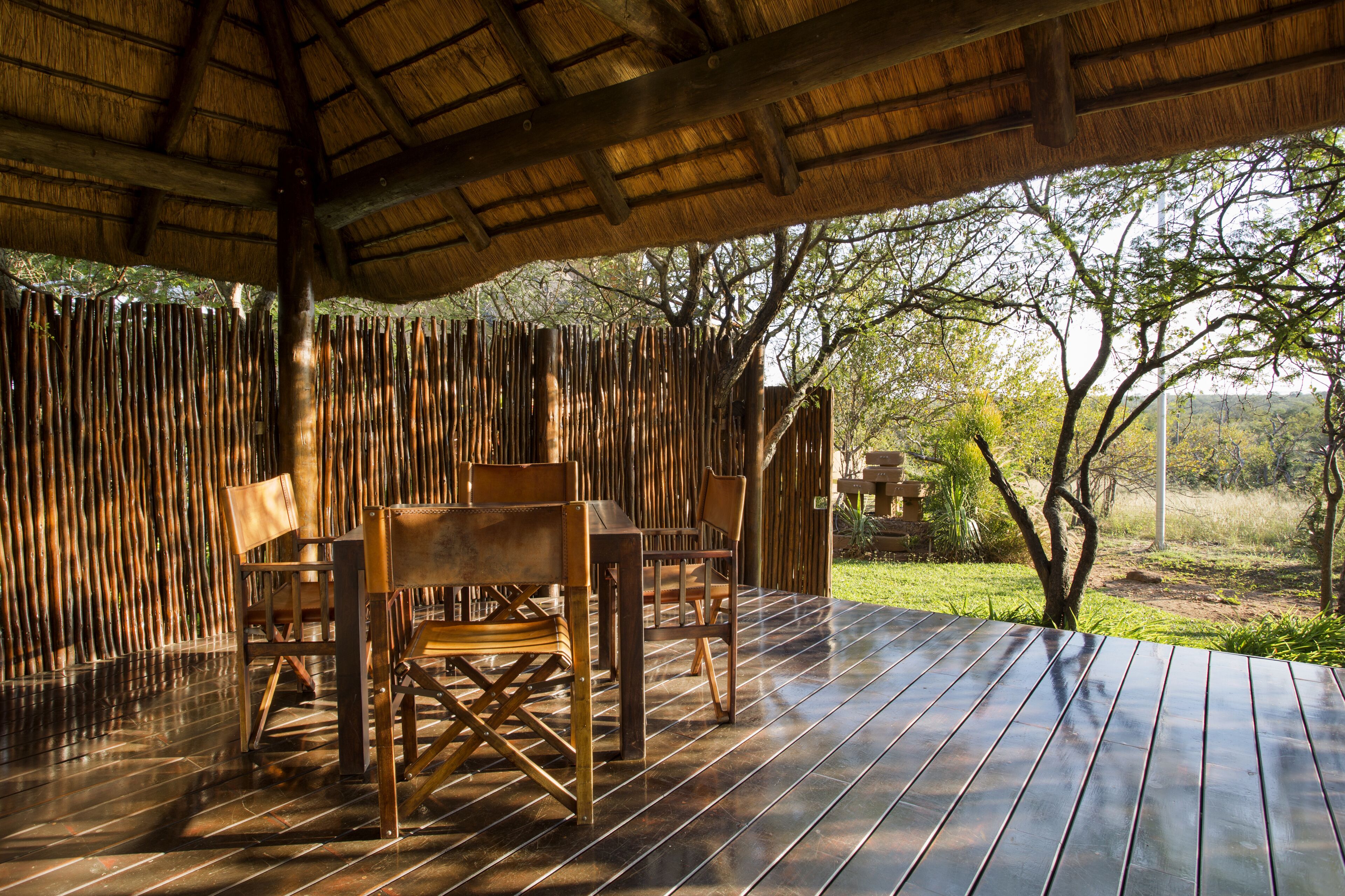 An outdoor dining area with a wooden table and chairs, surrounded by a bamboo fence, under a thatched roof.