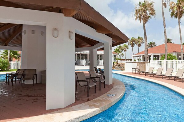 A covered pool area with lounge chairs and a view of palm trees and a building in the background.
