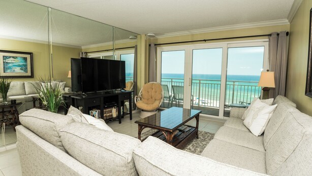 A spacious living room with a large flat-screen TV, comfortable seating, a coffee table, and a balcony with a view of the ocean.