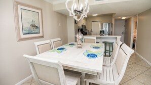 Comedor con una mesa blanca, sillas y una araña. Hay un cuadro en la pared y se ve una cocina al fondo.