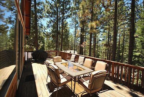 A wooden deck with a glass-top table and chairs, surrounded by tall pine trees.