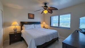Ein Schlafzimmer mit einem Bett, zwei Nachttischen mit Lampen, einem Deckenventilator, einem Fenster mit Jalousien und einem Schreibtisch mit einem Computermonitor.
