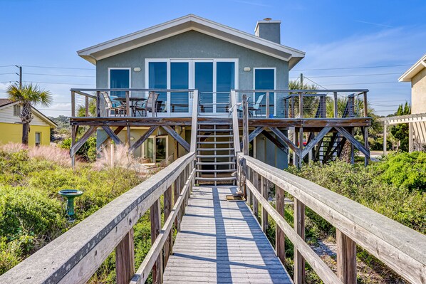 A house with a wooden deck leading to a glass door, surrounded by greenery. - Spacious living area, windows across entire length of home. (Fernandina Beach)