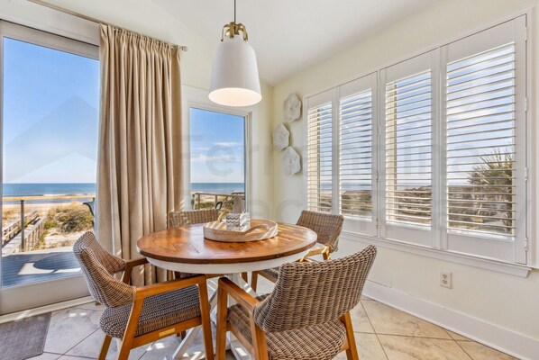 Un coin repas avec une table ronde en bois, des chaises en osier, un luminaire suspendu et une vue sur la plage grâce à de grandes fenêtres équipées de stores.