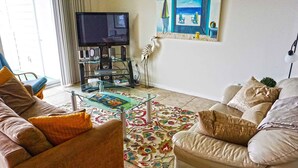A living room with a sofa, a coffee table, a TV, and a painting of a beach scene. - Thunder Island 128C-Baywater 39th St, Free WiFi, Pool, Elev, W/D, AC (Ocean City)