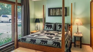 A bedroom with a four-poster bed, a bedside table with a lamp, and a view of the street through the window.