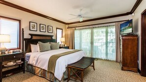 A hotel room with a large bed, a desk with a lamp, a small table, and a view of the outdoors through large windows.