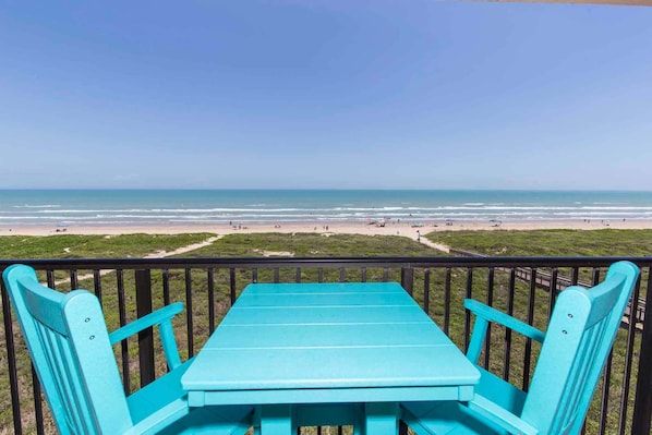 A turquoise table and chairs on a balcony overlooking a beach with a clear sky.
