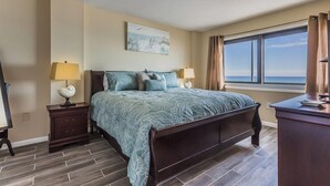 A bedroom with a bed, two bedside tables, a desk, a window with curtains, and a painting on the wall.