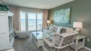 Un salon comprenant un canapé, des fauteuils, une table basse en verre et un fauteuil en osier. Un tableau est accroché au mur et, par la fenêtre, on aperçoit la plage.