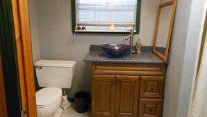 A compact bathroom with a toilet, a sink with a blue basin, a wooden vanity, a mirror, and a window with blinds.