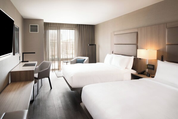 A hotel room with two beds, a desk, a chair, a TV, and a window with curtains.