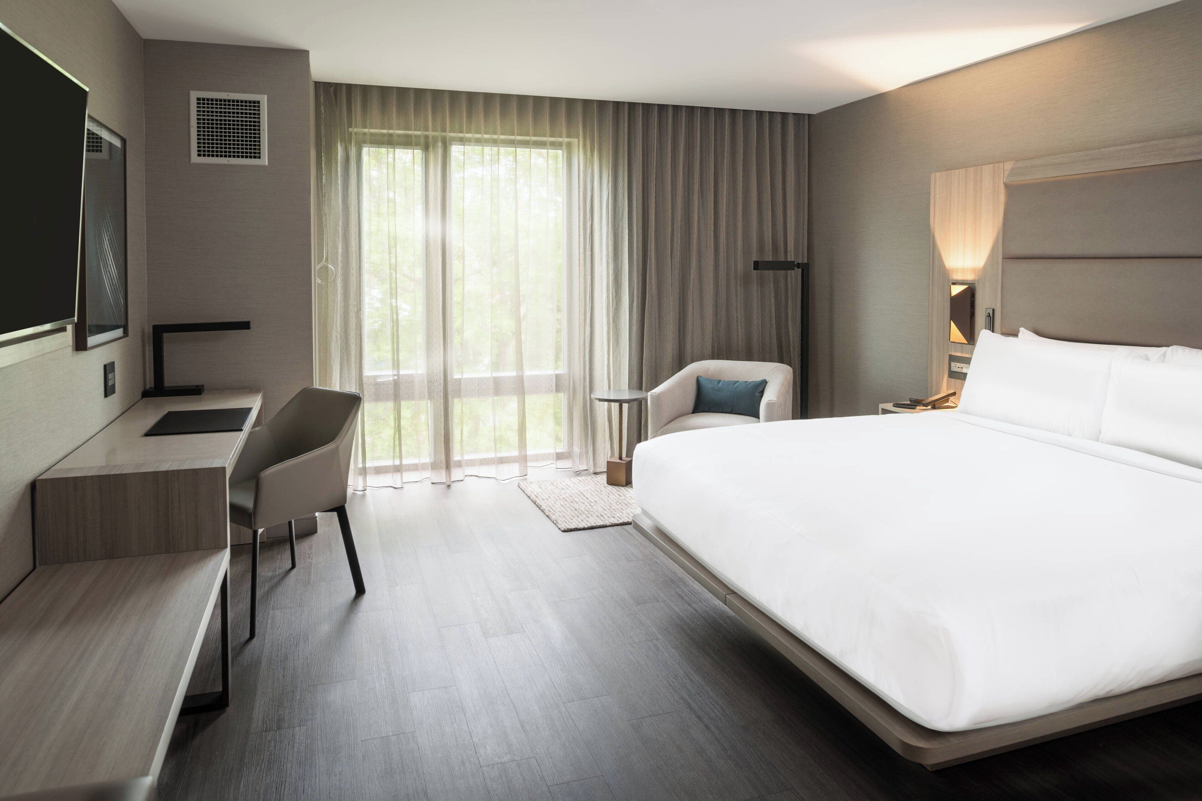 A modern hotel room with a large bed, a desk with a chair, a small table, and a TV.