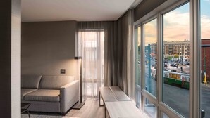 A modern hotel room with a sofa, bench, and large windows offering a city view. - AC Hotel by Marriott Boston Cleveland Circle (Boston)
