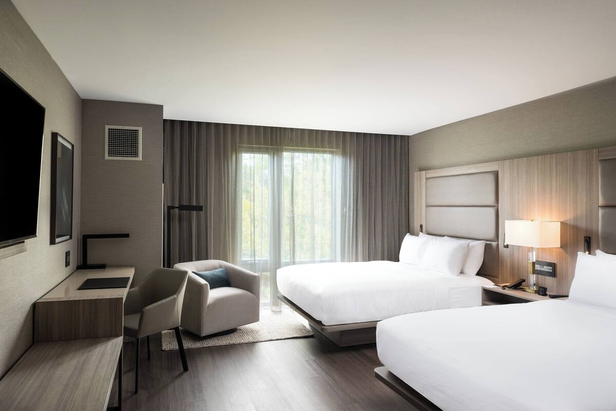 A hotel room with two beds, a desk, a chair, a TV, and a window with curtains.