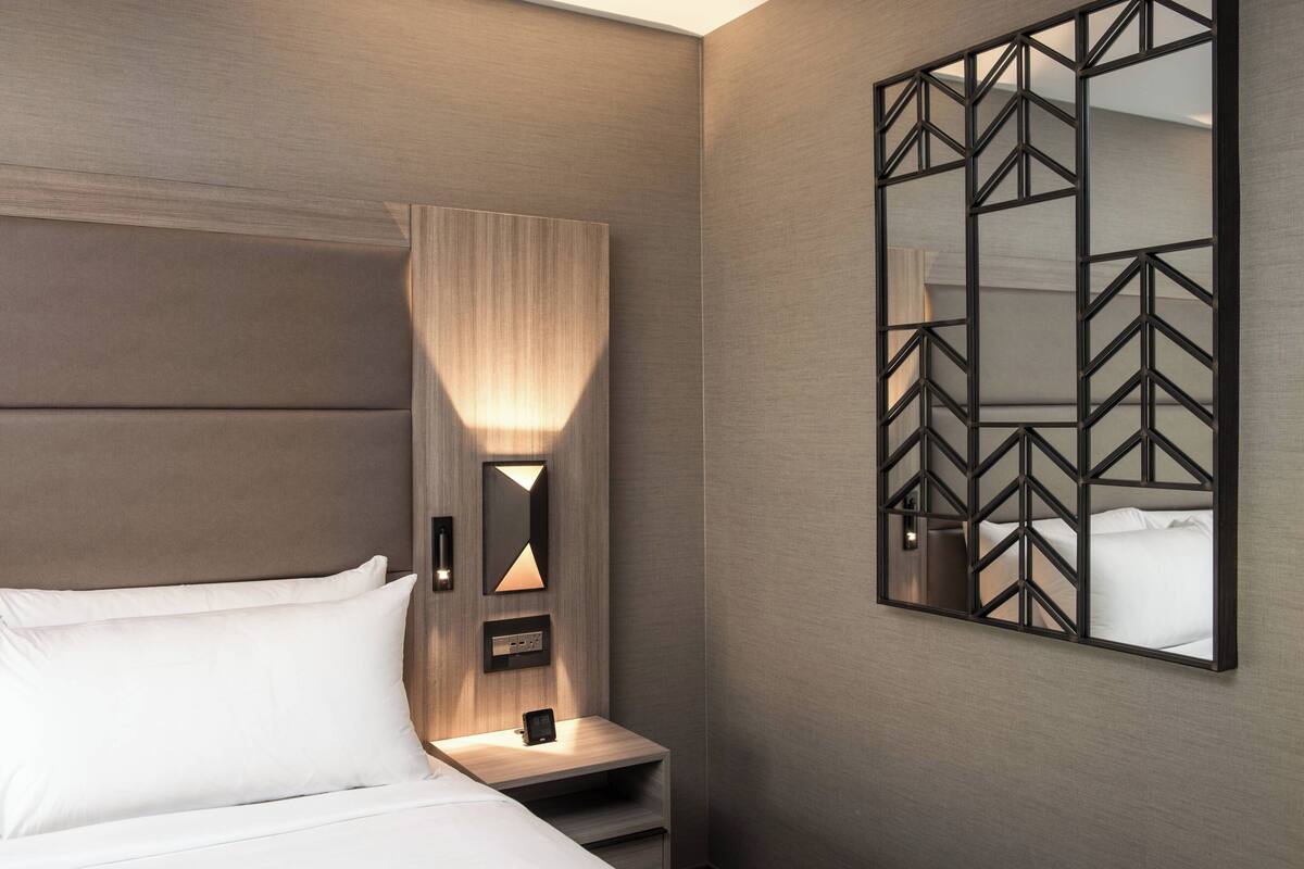 A modern hotel room with a large bed, a nightstand with a lamp, and a geometric mirror on the wall.