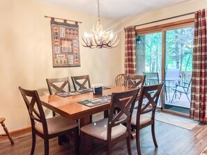A dining area with a wooden table and chairs, a chandelier, a wall tapestry, and a view of the outdoors through a glass door.