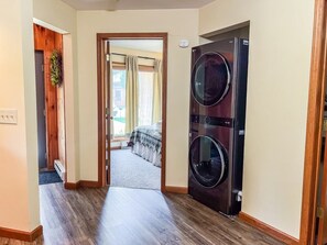 A laundry room with a double-door washer and dryer, a wooden door frame, and a carpeted floor leading to a bedroom with a bed and curtains.