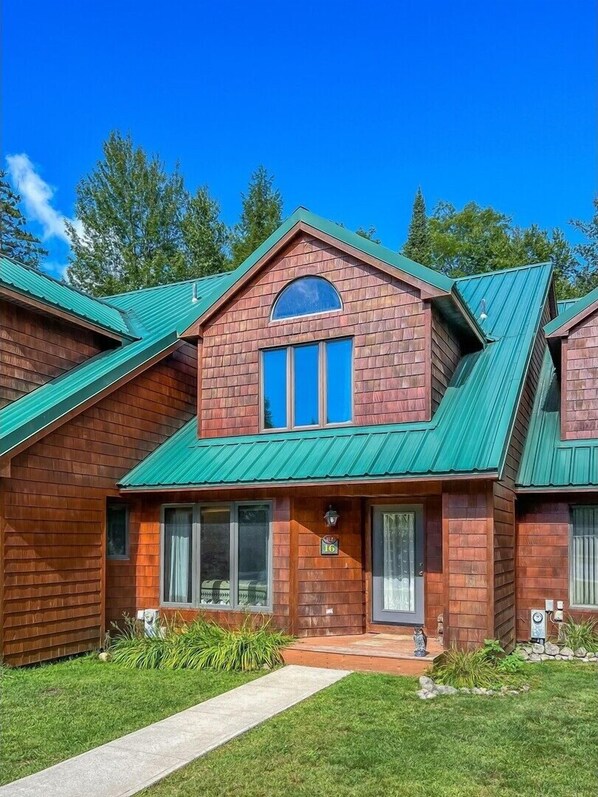 A two-story wooden house with a green metal roof, a front porch, and a small garden.