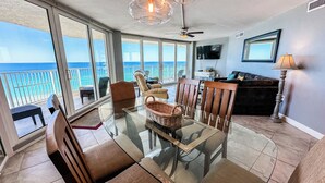 Un balcon spacieux offrant une vue dégagée sur l’océan, une table à manger avec un plateau en verre et des chaises, un panier en osier et un ventilateur de plafond.