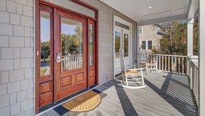 A wooden front door with a glass panel, a porch with a rocking chair, and a white railing. - 7 41st Avenue by AvantStay | Private Pool, Near Beach (Isle of Palms)